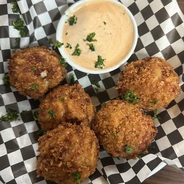 Five golden-brown Caribbean fritters with parsley, served with a creamy dipping sauce on checkered paper.