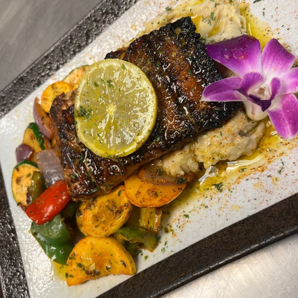 Grilled fish with a lemon slice, colorful vegetables, and a purple orchid on a white plate.