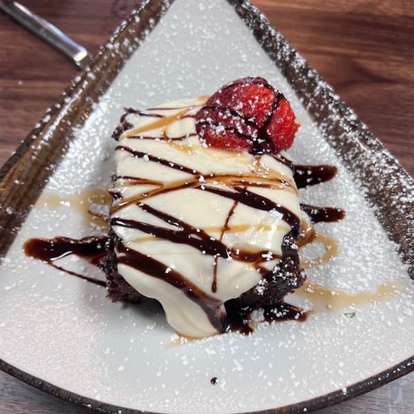 A chocolate brownie topped with white cream, chocolate, caramel, and a strawberry, dusted with powdered sugar.